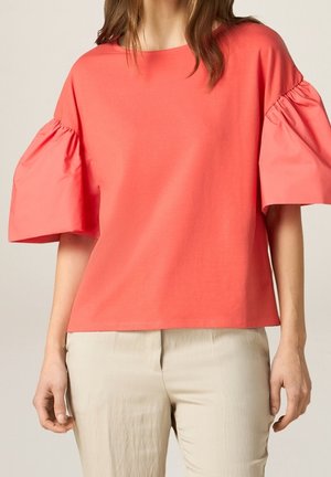 Frau trägt Korallenpinke Bluse mit kurzen Puffärmeln und hellbeige Hose, steht vor einem schlichten Hintergrund.