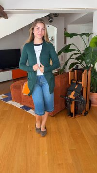 Žena stojaca v obývačke, oblečená v zelenom kardigáne, bielej košeli, modrých džínových šortkách a šedých balerínach, pri hnedej pohovke a izbových rastlinách.