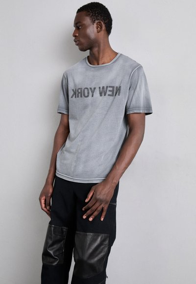 Graues Baumwoll-T-Shirt mit dem Aufdruck "NEW YORK" in fettem Schwarz, Rundhalsausschnitt und kurzen Ärmeln. Kombiniert mit schwarzen Hosen mit Lederakzenten.