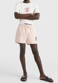 Bílá tričko s červeným logem "TOMMY JEANS", kombinované se světle růžovými pruhovanými šortkami s logem a černými sandály na klínku s detailním logem.