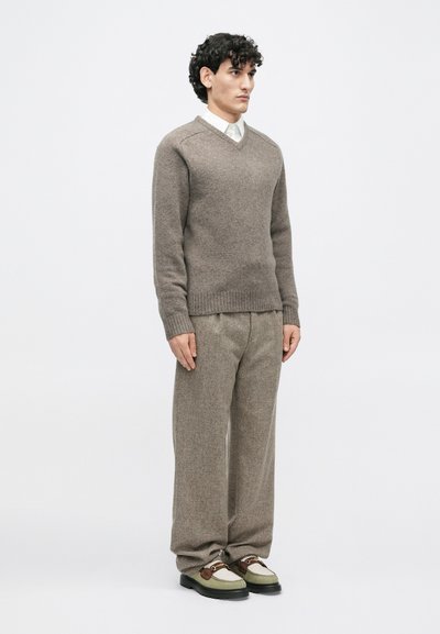 Maglione beige a V in morbido knit, abbinato a pantaloni beige a sacco con texture e scarpe con dettagli marroni e accenti verdi.