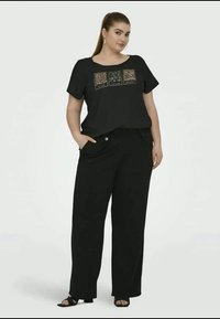 Top preto de manga curta com três padrões de estampas de animais em dourado e verde, combinado com calças pretas de corte solto e sandálias pretas de dedos abertos.