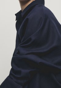 Chemise bleu marine à boutons avec col et coupe décontractée, présentant une texture lisse et un tissu fluide. Des détails subtils rehaussent le design.