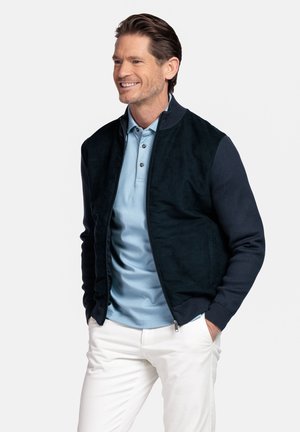 Man smiling, wearing a black zip-up jacket over a light blue polo shirt and white pants, hands in pockets, standing against plain background.