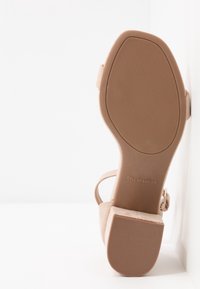 Semelle de sandale en daim beige avec talon carré, bride à la cheville et logo Call It Spring sur la voûte plantaire, sur fond blanc.