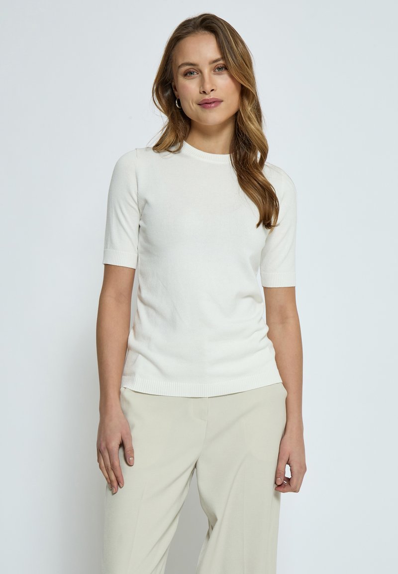 Lätt stickad kortärmad topp i off-white, med rund halsringning och ribbade kanter, parat med beiga byxor.