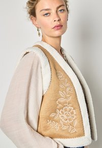 Braune Weste mit weißem Kunstpelzbesatz, mit floralem Stickerei. Der Stoff hat eine weiche Textur und eine ausgeprägte Kragenform.