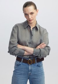Camicia a righe grigie con bottoni, maniche lunghe, abbinata a jeans azzurri a vita alta e una cintura marrone con una fibbia circolare dorata.