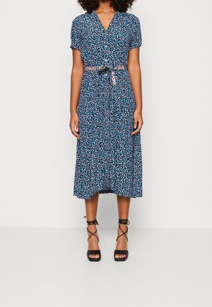 Kvinde iført en blå midikjole med blomsterprint og knapper samt et pink "Tommy Jeans" bælte, kombineret med sorte sandaler med hæl og stropper.