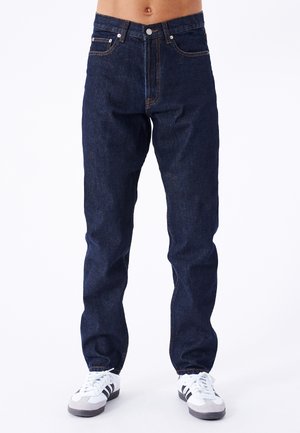 Jeans de mezclilla azul oscuro de corte recto, con costuras naranjas, cinco bolsillos y cierre frontal con botón. Llevados con zapatillas blancas.