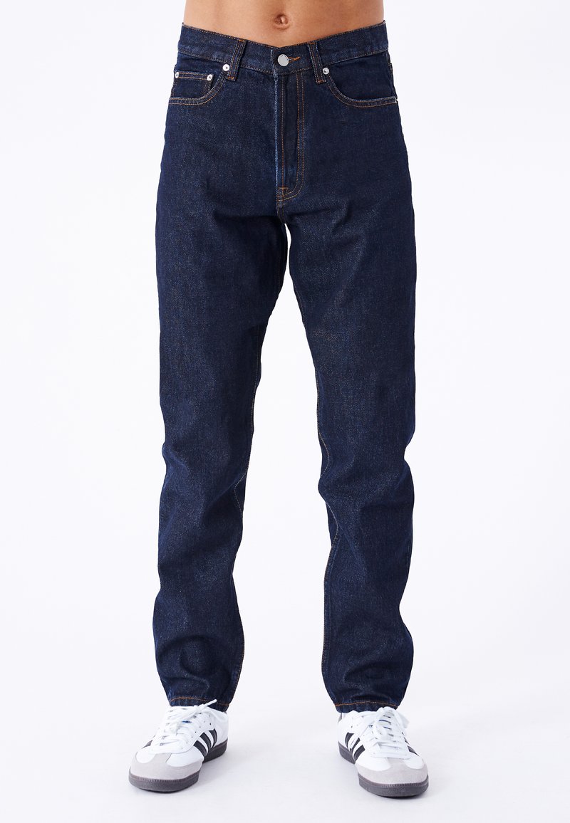 Jeans de mezclilla azul oscuro de corte recto, con costuras naranjas, cinco bolsillos y cierre frontal con botón. Llevados con zapatillas blancas.