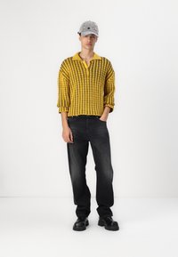Pull en tricot rayé jaune avec un col, associé à un jean noir et des chaussures noires imposantes. Une casquette grise avec des accents colorés complète le look.