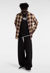 Camisa de cuadros en púrpura y amarillo sobre una camiseta negra, combinada con pantalones holgados negros y zapatillas negras con suelas blancas.