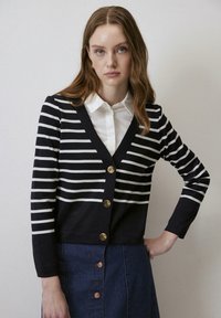 Marineblauwe en witte gestreepte cardigan met een V-hals, voorzien van gouden knopen en lange mouwen, gecombineerd met een spijkerrok.