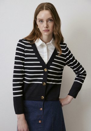 Cardigan rayé bleu marine et blanc avec un col en V, doté de boutons dorés et de manches longues, associé à une jupe en denim.