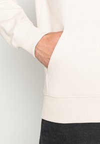 Sudadera color crema hecha de material suave, con puños y dobladillo acanalados, y un bolsillo lateral visible a la derecha.