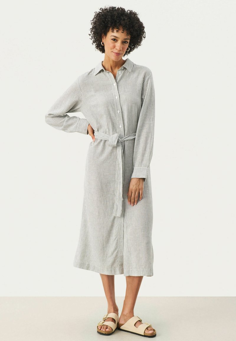 Abito a camicia a righe con maniche lunghe in grigio chiaro e bianco, con chiusura a bottoni, vita legata e vestibilità comoda. Abbinato a sandali neutri.