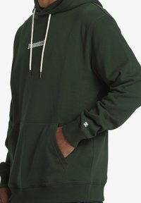Felpa verde realizzata in tessuto misto cotone, con tasca frontale a marsupio, cappuccio regolabile con cordino e logo sulla manica sinistra.