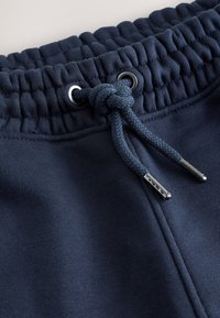 A tengerészkék melegítőnadrág húzott derékpánttal, fém karikákkal, húzózsinórral és sima, egyszerű mintával rendelkezik.