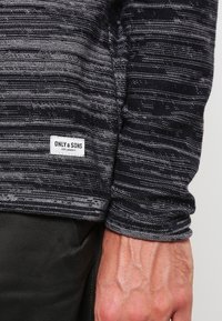 Chemise noire et grise à manches longues avec un motif de rayures horizontales, texture douce, et une étiquette portant la mention "ONLY & SONS" à l'ourlet.