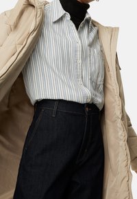 Abrigo acolchado beige sobre una camisa de rayas blanca y azules, combinado con jeans de mezclilla oscuros, que presenta un diseño clásico de botones y bolsillos frontales.