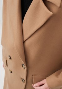Cappotto doppiopetto in morbido tessuto beige. Presenta grandi bottoni frontali, rever a punta e una tasca laterale, evidenziando un design elegante.