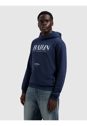 Jonge man in een marineblauwe hoodie met witte tekst "BALLIN", staand tegen een effen lichtgrijze achtergrond, met lichtblauwe jeans aan.