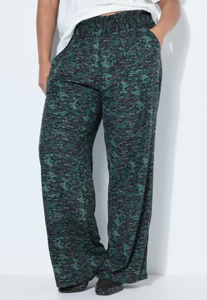 Persona con pantalones holgados de camuflaje en verde y negro, con las manos en los bolsillos, combinados con zapatos negros texturizados.