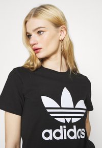 Mustmust must kaelusega ja lühikeste varrukatega must puuvillane T-särk, millel on suur valge Adidas trefoil logo ja kaubamärgi nimi.