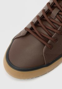 Brązowe skórzane trampki o teksturowanym wykończeniu, z brązowymi sznurówkami, gumową podeszwą oraz czarną obręczą wokół noska.