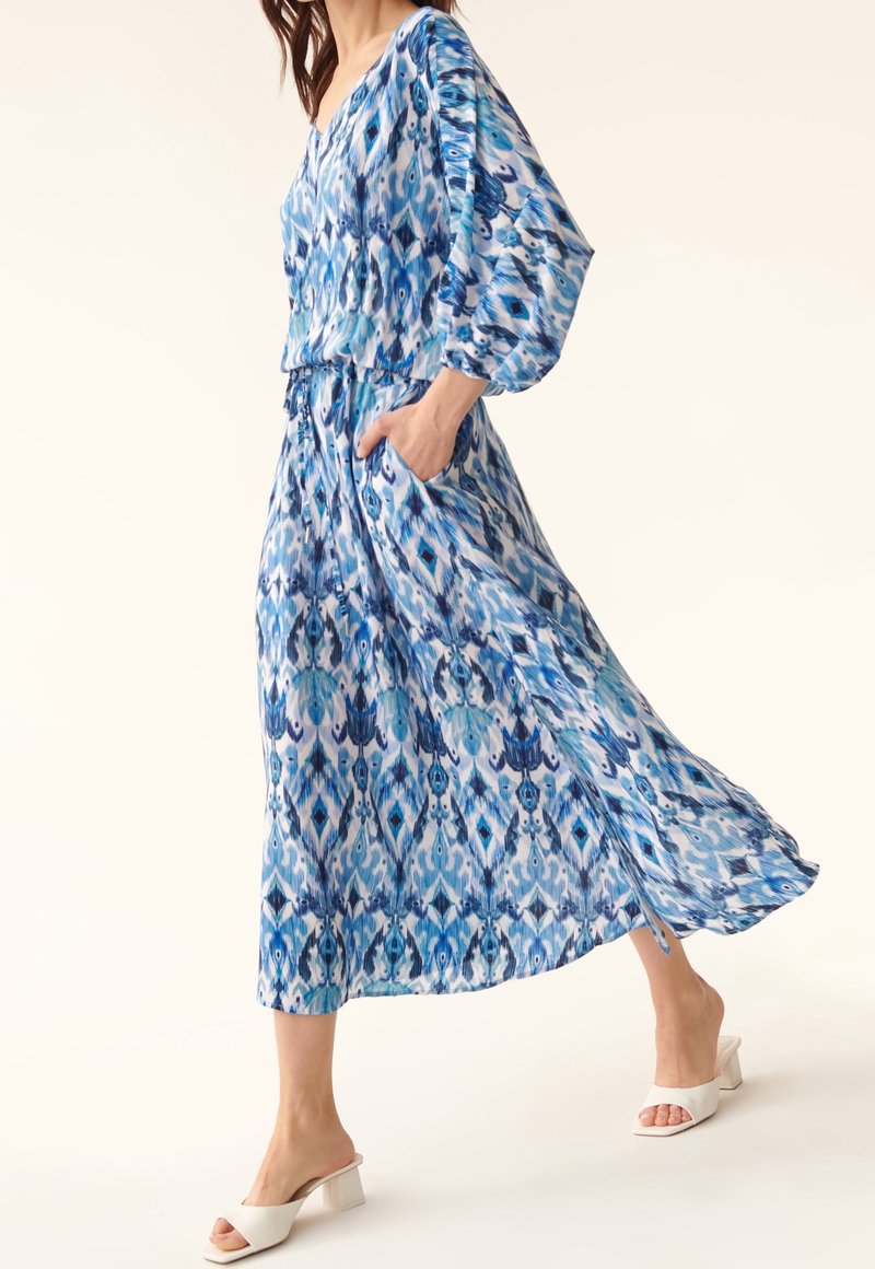Femme marchant dans une robe midi fluide bleue à motifs avec des manches longues et des sandales blanches à talons, la main dans la poche de la robe.