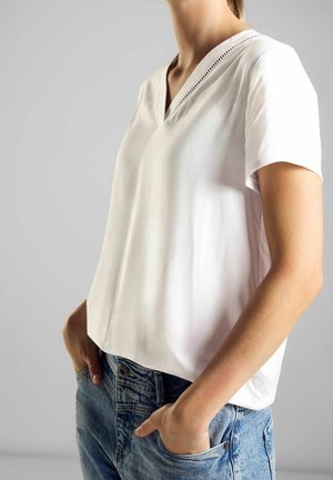 Top blanc à manches courtes avec un col en V et des détails raffinés au niveau du col, associé à un jean en denim bleu clair. Texture lisse.