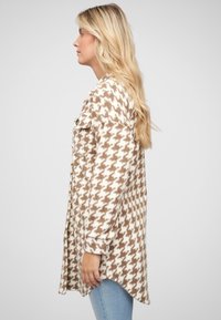 Jachetă lungă cu model houndstooth în nuanțe de maroniu și crem, cu guler, buzunare laterale și tiv curbat; realizată dintr-un material moale și texturat.