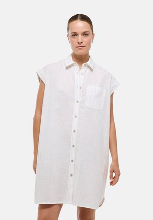 Femme aux cheveux attachés portant une robe blanche sans manches boutonnée avec une poche poitrine, debout devant un fond uni.