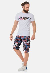 Weißes T-Shirt mit kurzen Ärmeln und mehrfarbigem Text; blaue, rote und schwarze Cargo-Shorts mit abstraktem Muster und dem Text "POWER". Weiße Sportschuhe.