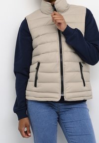 Beige gewatteerd vest met hoge kraag, ritssluiting aan de voorkant en zwarte accenten. Draagt over een marineblauwe sweatshirt, voorzien van twee ritssluitingen zakken.