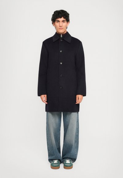 Man in een zwart knopenjas, wijde lichtblauwe jeans en groene sneakers voor een egale witte achtergrond.