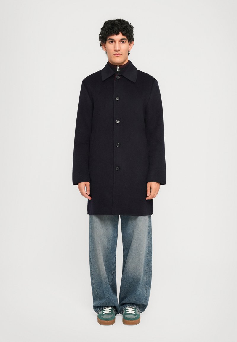 Man standing in a black buttoned coat, wide light blue jeans, and green sneakers against a plain white background.