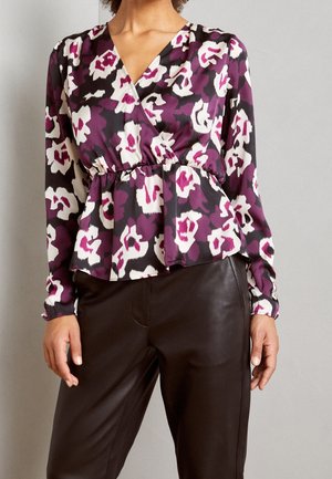 Frau trägt eine langärmelige Bluse mit lila, weißen und schwarzen Blumen, V-Ausschnitt und schwarze Lederhose, steht vor einem einfarbigen Hintergrund.