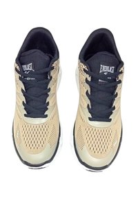 Scarpe da ginnastica beige con tomaia in rete traspirante, lacci neri e dettagli con logo; presentano una suola imbottita e superficie testurizzata.