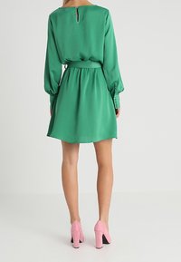Grünes, langärmliges Kleid mit tailliertem Schnitt, fließendem Rock und Rückendetail mit Schlüssel-Ausschnitt, kombiniert mit rosa High Heels.