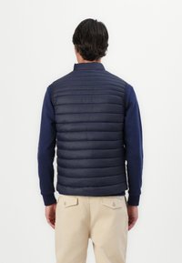 Vesta puffy de culoare bleumarin, cu un design matlasat, guler înalt și buzunare laterale, purtată peste o cămașă cu mânecă lungă, asortată cu pantaloni de culoare deschisă.