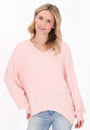 Lyserød waffle-strikket pullover med V-hals, brede ærmer og buet kant. Har en struktureret overflade og løs pasform. Synlige denim jeans.