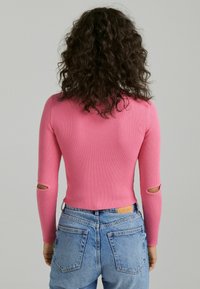Fitted ribbed sweater in vibrant pink with long sleeves featuring cutouts. Paired with high-waisted blue denim jeans and a simple waistband label.