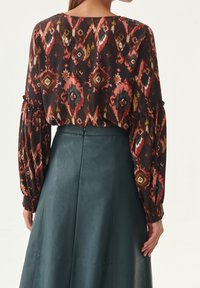 Blusa a maniche lunghe realizzata in un tessuto scuro e fantasioso con forme geometriche in rosso, arancione e verde, abbinata a una gonna in pelle verde.
