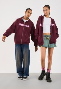 Deux personnes avec des cheveux tressés portant des sweat-shirts assortis de couleur bordeaux, l'une en jean ample et baskets, l'autre en jupe en denim, top court blanc et bottes.