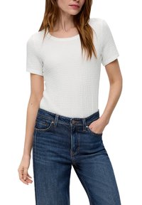Biała, strukturalna bluzka z krótkim rękawem w połączeniu z ciemnoniebieskimi dżinsami. Bluzka ma okrągły dekolt i dopasowany fason.
