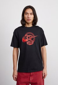 Juodas medvilninis marškinėlis su dideliu raudonu Nike logotipu, apipintu gyvačių dizainu, apvaliu kaklu ir trumpomis rankovėmis.