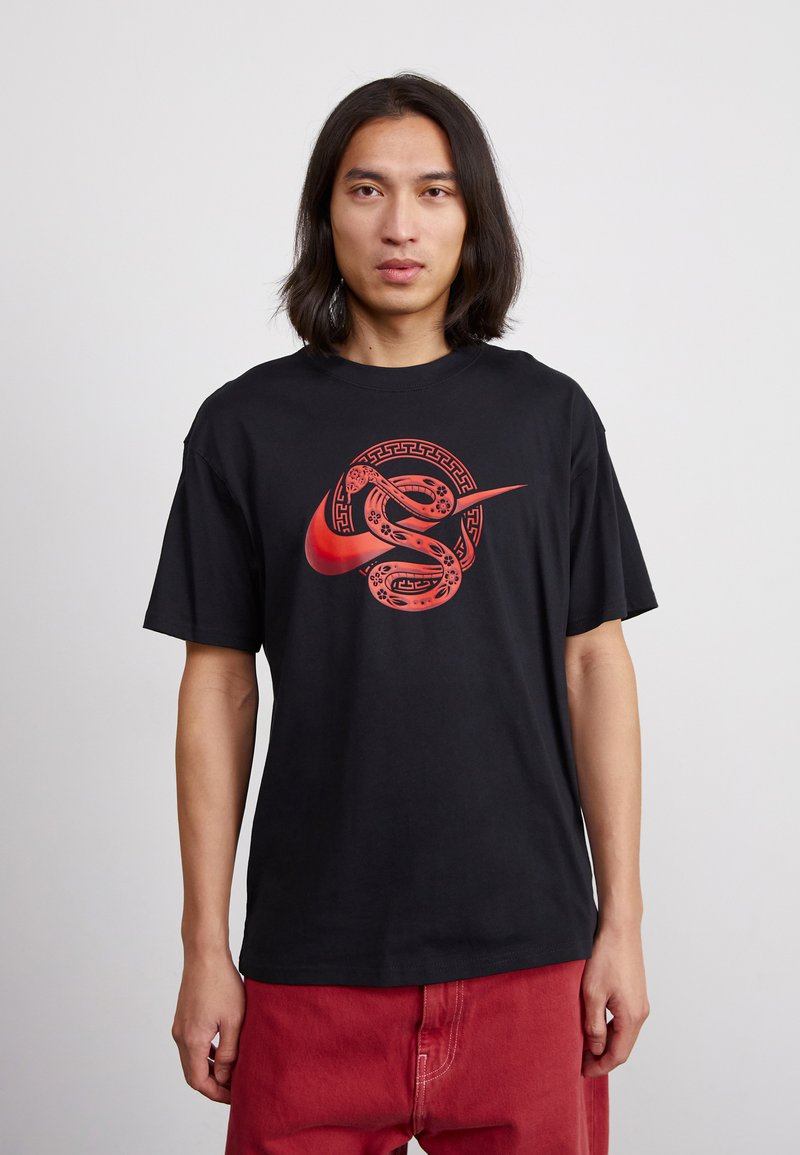 Juodas medvilninis marškinėlis su dideliu raudonu Nike logotipu, apipintu gyvačių dizainu, apvaliu kaklu ir trumpomis rankovėmis.