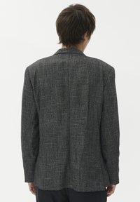 Mees, seljas tumehall struktureeritud bleiser, seisab seljaga seina vastu, mille taust on ühtlane heledavärviline.